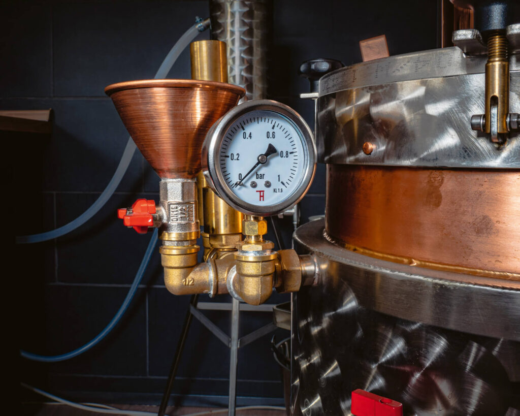 Close-up of a copper funnel, brass valves, and round pressure gauge attached to a stainless steel and copper gin still inside a distillery.