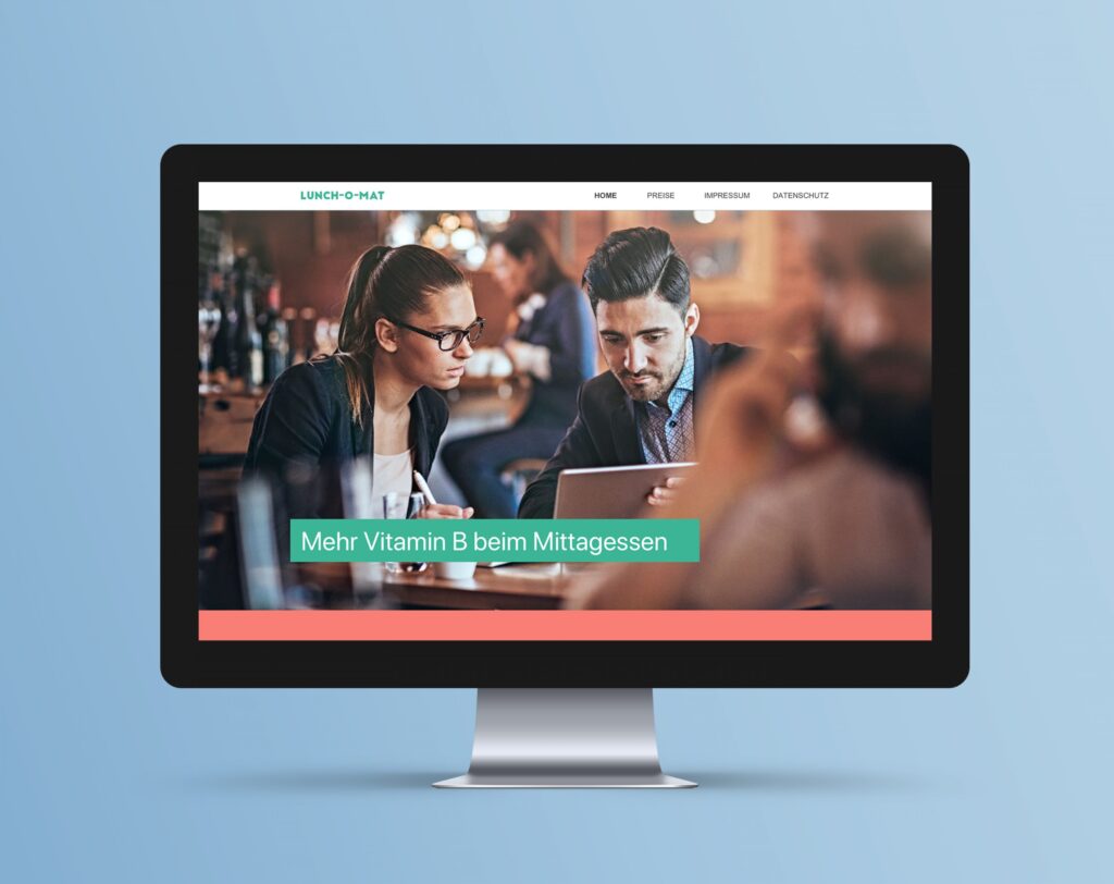 A desktop monitor displaying the "Lunch-O-Mat" website, featuring a photo of two colleagues looking at a tablet and the text "Mehr Vitamin B beim Mittagessen".