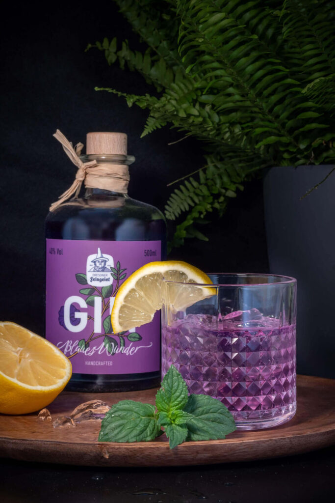 Bottle of Dresdner Feingeist GIN Blaues Wunder with a faceted glass of purple gin cocktail, garnished with a lemon slice, fresh mint leaves, and a halved lemon on a wooden board against a dark background with green fern leaves.