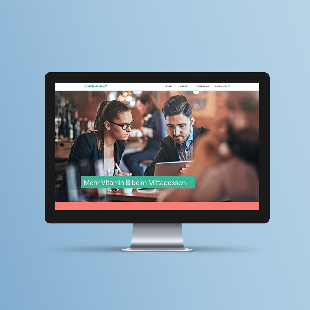 Monitor displaying the "Lunch-o-mat" website featuring a photo of two professionals looking at a tablet in a restaurant with the headline "Mehr Vitamin B beim Mittagessen."