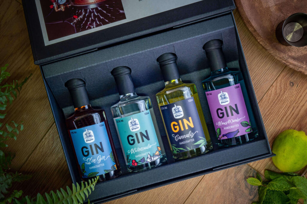 Gift box with four Dresdner Feingeist gin bottles—Sloe Gin, Blütenzauber, Cannabis, and Blütenwunder—neatly arranged in a dark foam insert on a wooden surface with green foliage accents.