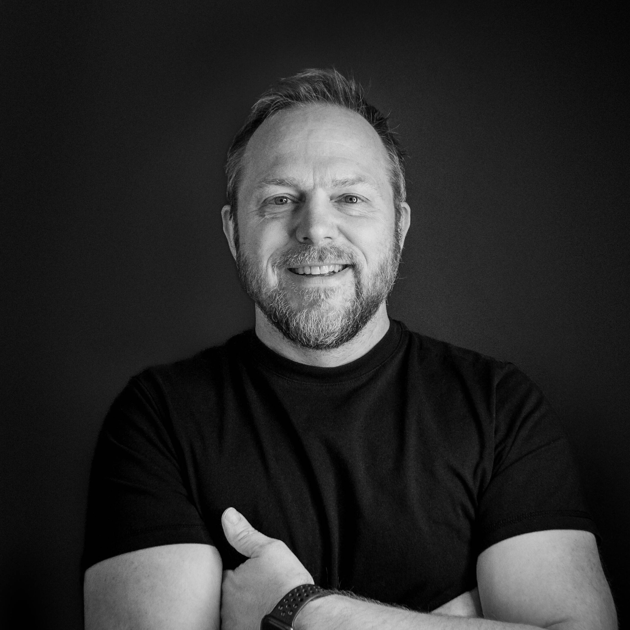 Black and white portrait of a smiling man with a beard and short hair, wearing a dark t-shirt and crossing his arms.